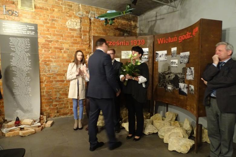 Uczniowie, nauczycielka z bukietem kwiatów i mężczyzna na tle wystawy ze zdjęciami