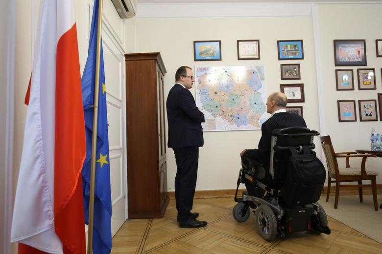 Mężczyzna na wózku i męzczyzna stojący przed mapą polski w gabinecie, gdzie w rogu stoją flagi Polski i Unii