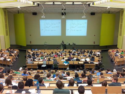 Pełna sala wykładowa. Przy pulpicie stoi mężczyzna prowadzący wykład, a nad nim wyświetlane są slajdy prezentacji przedstawiające równania matematyczne.