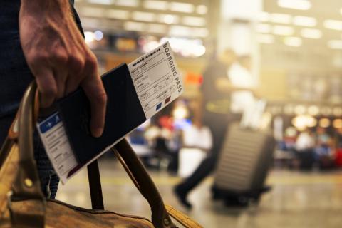 Na pierwszym planie dłoń trzymająca paszport i kartę pokładową, w tle hala lotniska