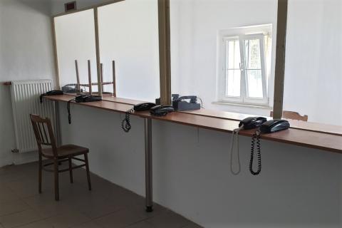 Pomieszczenie przedzielone ścianą z oknem z pleksi z krzesłami i telefonami po obu stronach szyby