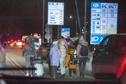 Grupa kilku osób z dziećmi, z torbami i plecakami, idzie nocą między samochodami. Na drugim planie znaki drogowe informujące o wjeździe do Polski