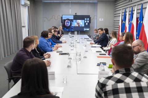 Ludzie siedzący przy długim stole, w tle ekran ze zdjęciami ludzi, po prawej flagi Polski i Unii Europejskiej