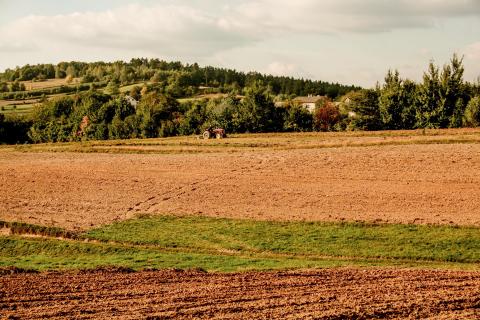 Krajobraz rolniczy - rolnik pracujący na ciągniku w polu, za którym widać zarośla przysłaniające wiejską zabudowę.