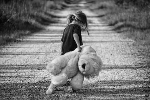 Czarno-białe zdjęcie smutnej dziewczynki idącej drogą i ciągnącej dużego, pluszowego misia