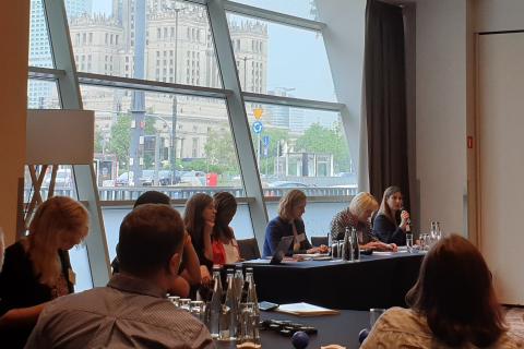 Cztery osoby siedzą za stołem prezydialnym, jedna z nich wypowiada się do mikrofonu. W tle okno, przez które widać centrum Warszawy.