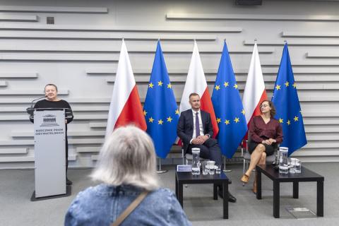 Trzy osoby na tle flag Polski i UE. Jedna stoi za mównicą i przemawia, dwie siedzą na krzesłach i słuchają wystąpienia