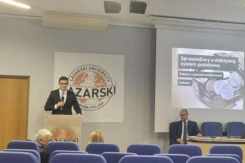 ZRPO Valeri Vachec wypowiada się przy mównicy w sali wykładowej. Na ścianie za nim widoczne jest logo uczelni Łazarskiego i slajd z prezentacji