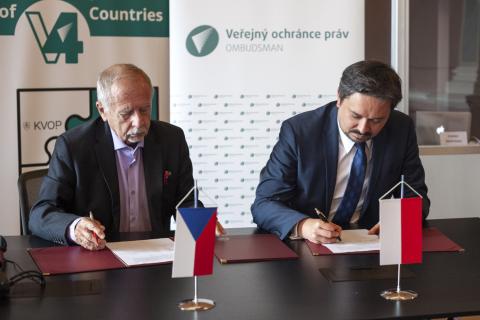 Dwie osoby siedzące za stołem podpisują dokument, na stole na stojakach flagi Polski i Czech