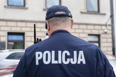 Stojąca osoba w mundurze i czapce, na plecach napis POLICJA
