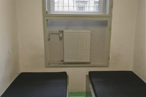 dwa materace, kaloryfer i zakratowane okno w policyjnym pomieszczeniu dla zatrzymanych 