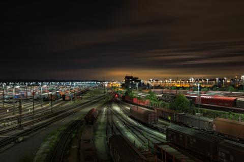 Nocna panorama jasno oświetlonej bocznicy kolejowej. Na horyzoncie miasto, a nad nim pomarańczowa łuna oświetlenia