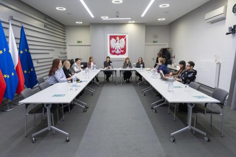 Uczestnicy warsztatów siedzą przy konferencyjnym stole w kształcie podkowy. W tle godło Polski oraz flagi Polski i Unii Europejskiej