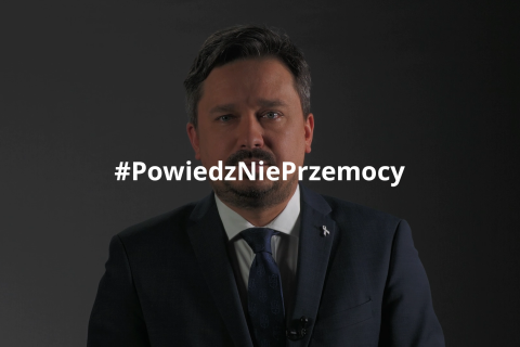 RPO Marcin Wiącek stoi na ciemnym tle. Na środek zdjęcia naniesiony jest napis hasztag "Powiedz Nie Przemocy"