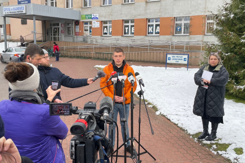 Zastępca RPO Wojciech Brzozowski (w centrum kadru) wypowiada się do mikrofonów stojących przed nim dziennikarzy. Na pierwszym planie jest kamera telewizyjna. W tle jest budynek wielopiętrowy z zadaszonym wejściem, na którym znajduje się szyld z napisem "Szpital w Hajnówce im. lek. Włodzimierza Mantiuka"