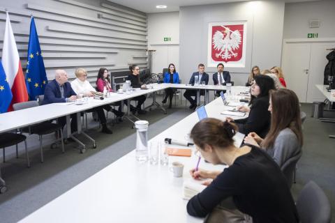 Grupa kilkunastu osób siedzi przy dużym stole konferencyjnym, w tle godło Polski i flagi Polski i UE