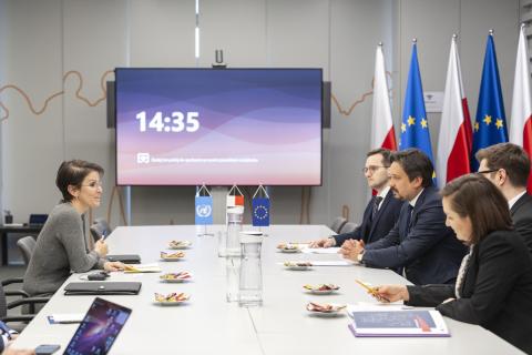 Siedem osób siedzi po obu stronach stołu i rozmawia. W tle stojące na środku stołu małe flagi Polski, UE i ONZ