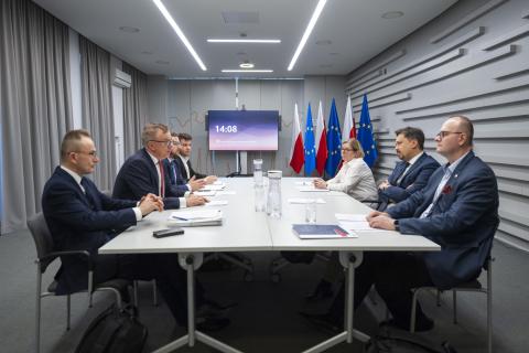 Siedem osób siedzi po obu stronach stołu i rozmawia. W tle flagi Polski i UE