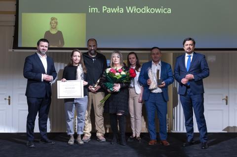 Siedem osób, w tym RPO Marcin Wiącek i laureaci Nagrody, pozują do zdjęcia na podium. Stojąca w centrum kobieta trzyma kwiaty, a stojąca po lewej stronie kobieta trzyma tabliczkę z nekrologiem.