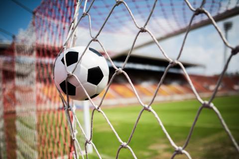 zdjęcie przedstawiające piłkę do piłki nożnej wpadającą do bramki, w tle zielone boisko i trybuny