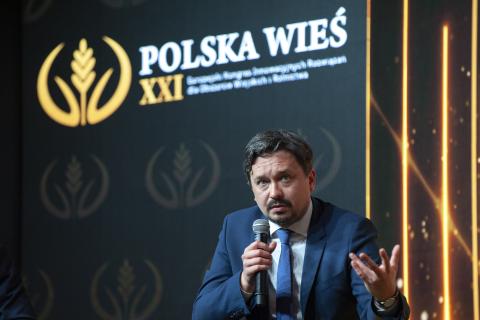 RPO Marcin Wiącek siedząc mówi do mikrofonu. Za nim wyświetla się logo konferencji.