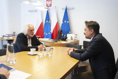 Dwie osoby siedzą przy stole i rozmawiają, w tle flagi Polski i UE