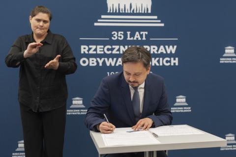 RPO Marcin Wiącek siedzi za stołem i podpisuje dokument, za nim stoi tłumacz polskiego języka migowego