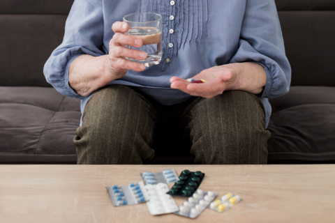 seniorka siedzi w jednej ręce trzyma szklankę z wodą a w drugiej leki 