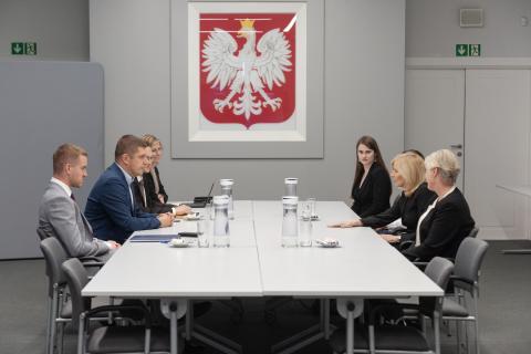 Osiem osób siedzi po dwóch stronach dużego prostokątnego stołu w sali konferencyjnej, w tle na ścianie godło polski - biały orzeł w koronie na czerwonym polu