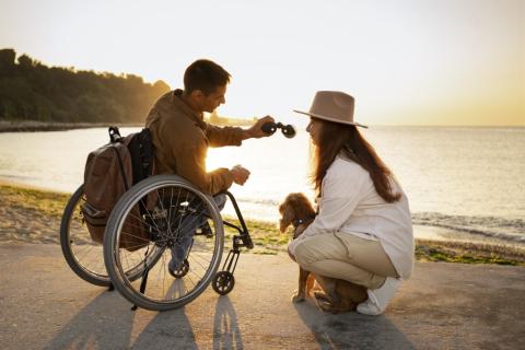 osoba na wózku na plaży przy zachodzie słońca podaje lornetkę kobiecie z  psem
