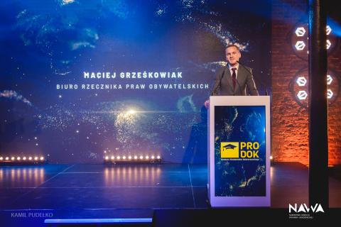 Osoba przemawiająca za mównicą na scenie, w tle wyświetlacz z napisem "Maciej Grześkowiak, Biuro Rzecznika Praw Obywatelskich"