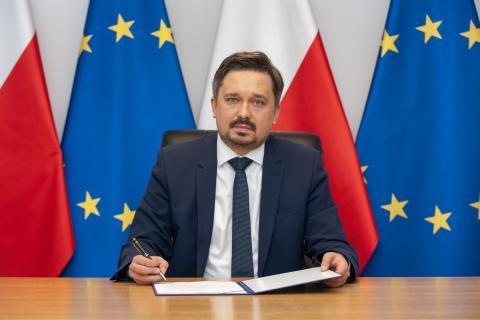 RPO Marcin Wiącek siedzi za biurkiem na tle flag Polski i UE, w dłoni trzyma pióro nad kartką papieru
