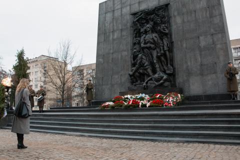 Anna Grzelak stoi z pochyloną głową pod Pomnikiem Bohaterów Getta, pod którym leżą wieńce. Przy pomniku wartę pełni dwóch żołnierzy