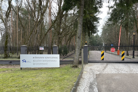 Brama w ogrodzeniu ze szlabanem w lesie, a obok na tablicy napis "Ośrodek szkolenia Służby Więziennej w Popowie"