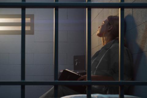 kobieta siedzi oparta o ścianę w celi więziennej widziana zza krat 