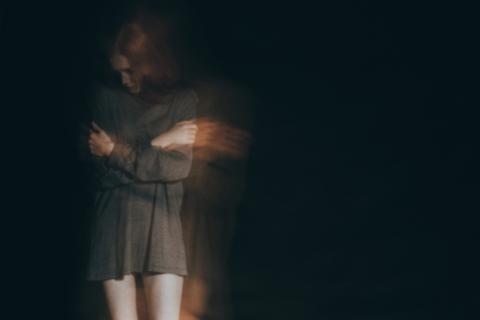 kobieta w rozciągniętej koszuli stojąca w ciemnym pomieszczeniu z pochyloną głową i obejmująca się rękami