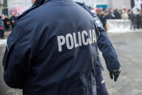 Funkcjonariusz policji w mundurze z napisem "POLICJA" na plecach, a w oddali tłum ludzi