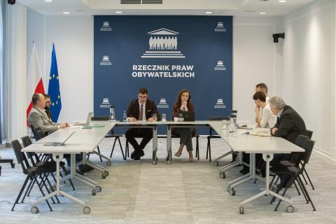 Siedmioro uczestników spotkania siedzi przy konferencyjnym stole. W tle flagi Polski i Unii Europejskiej