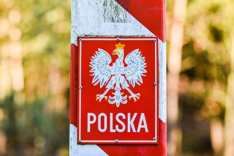 Biało-czerwony słup graniczny, na którym przybita jest tabliczka z godłem Polski i napisem "POLSKA" na leśnym tle