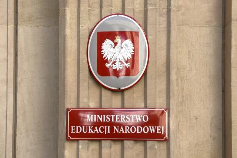 Czerwona tablica z białym napisem "Ministerstwo Edukacji Narodowej" powyżej owalna tablica z godłem Polski - białym orłem w koronie na czerwonym polu