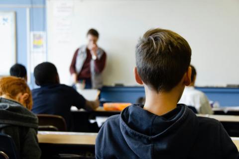 Dziecko w klasie widziane od tyłu, w tle prowadzący zajęcia oraz tablica szkolna