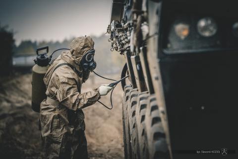Żołnierz w kombinezonie chemicznym i masce przeciwgazowej czyści ze skażeń chemicznych transporter opancerzony