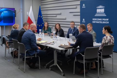 Dziewięć osób siedzi przy dużym prostokątnym stole w sali konferencyjnej, w tle na ścianie napis "Rzecznik Praw Obywatelskich" oraz flagi Polski i UE