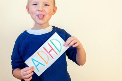 Uśmiechnięte dziecko wystawia język trzymając w dłoniach kartkę z napisem ADHD