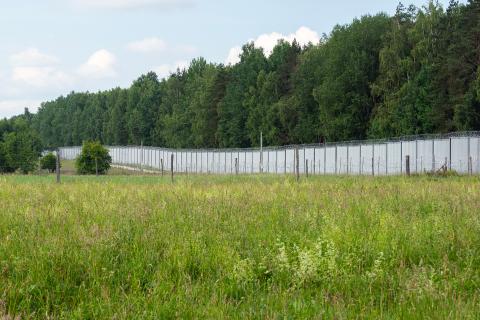 Ciągnący się wzdłuż lasu stalowy mur graniczny zakończony zwojami drutu kolczastego. Przez murem z polskiej strony granicy znajdują się dalsze zasieki.
