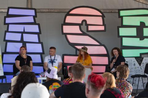 Uczestnicy panelu siedzą na scenie namiotu ASP. Na pierwszym planie siedzą słuchający ich uczestnicy festiwalu