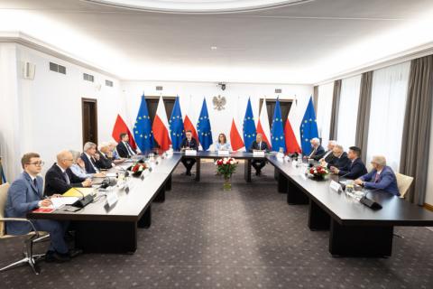 Kilkanaście osób siedzi przy stołach ustawionych w podkowę, za stołami przy ścianie rząd flag Polski i UE