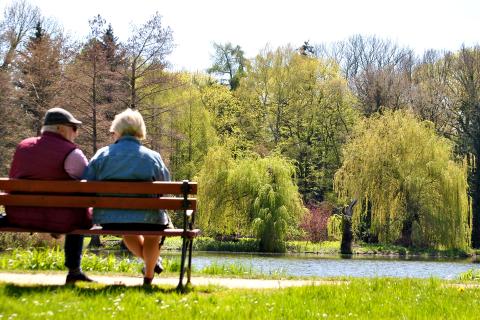 Mężczyzna i kobieta, oboje w starszym wieku, siedzą na ławce przed stawem w parku i rozmawiają 