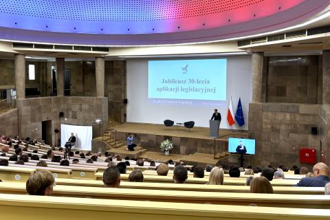 Kilkadziesiąt osób siedzi w dużej auli wykładowej i słucha wystąpienia osoby mówiącej na scenie. Za sceną, na dużym ekranie napis "Jubileusz 30-lecia aplikacji legislacyjnej"