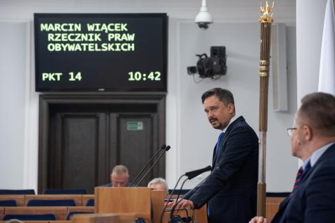 RPO Marcin Wiącek stoi za mównica na sali plenarnej Senatu RP i przemawia, na ścianie w oddali wyświetlacz z tekstem "Marcin Wiącek, Rzecznik Praw Obywatelskich"